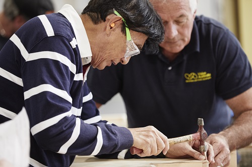 A man hammers a nail into a piece of wood under the supervision of another man