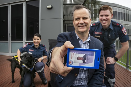 Chris holds a braille Ambulance Victoria ID card and stands with his Seeing Eye Dog Odie and Paramedics Kate and Carl