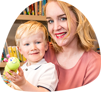 A woman with long blonde hair has her toddler son on her lap. They are both smiling at the camera.