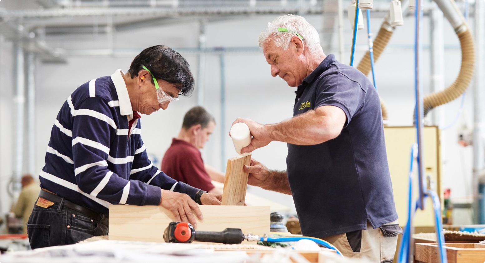 Two men assisted by a Vision Australia employee, wearing safety goggles work together in a woodworking shop. The employee applies glue to a wooden joint while one of the men holds the pieces in place.