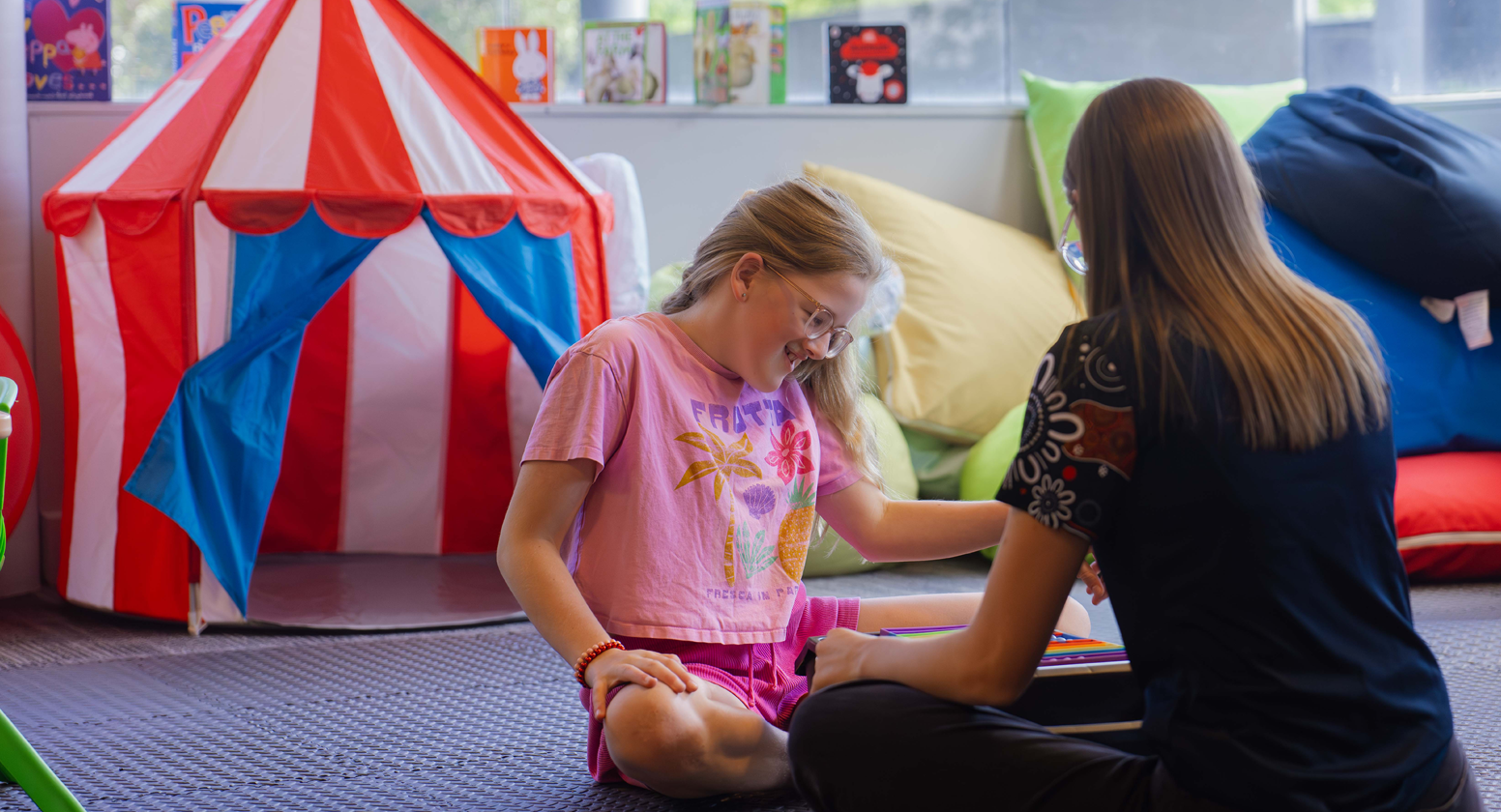 Young girl and Vision Australia team member are seated on the ground opposite each other. In the background there is a circus play tent, toys and books.