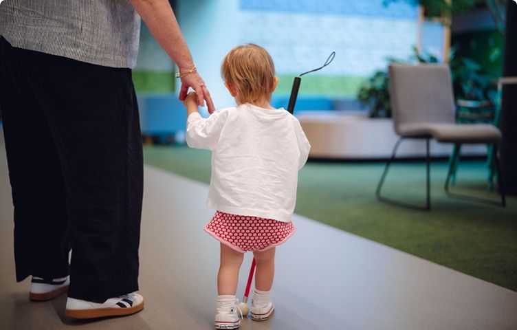 A young child is walking, holding a white cane and a Vision Australia team member's hand.
