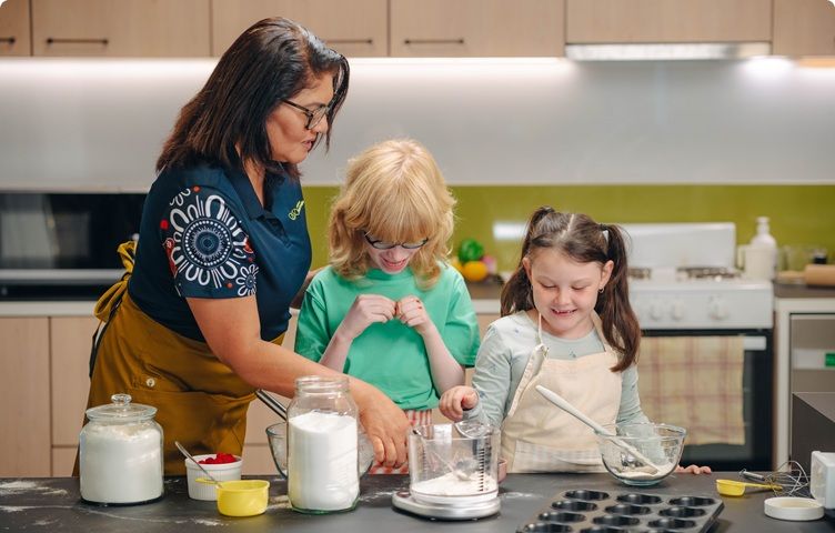 A Vision Australia team member is helping two children cooking in a kitchen.