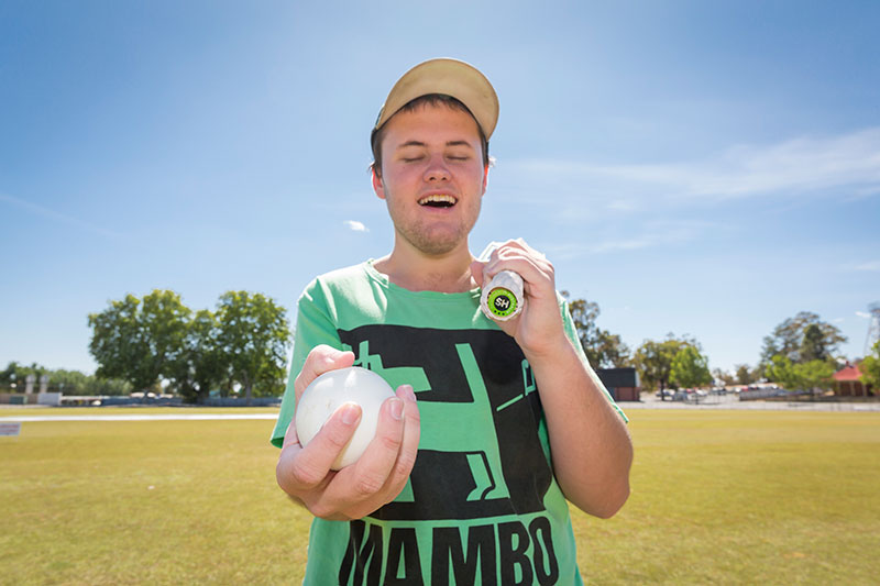 Zac stands on cricket oval and holds a cricket bat and ball