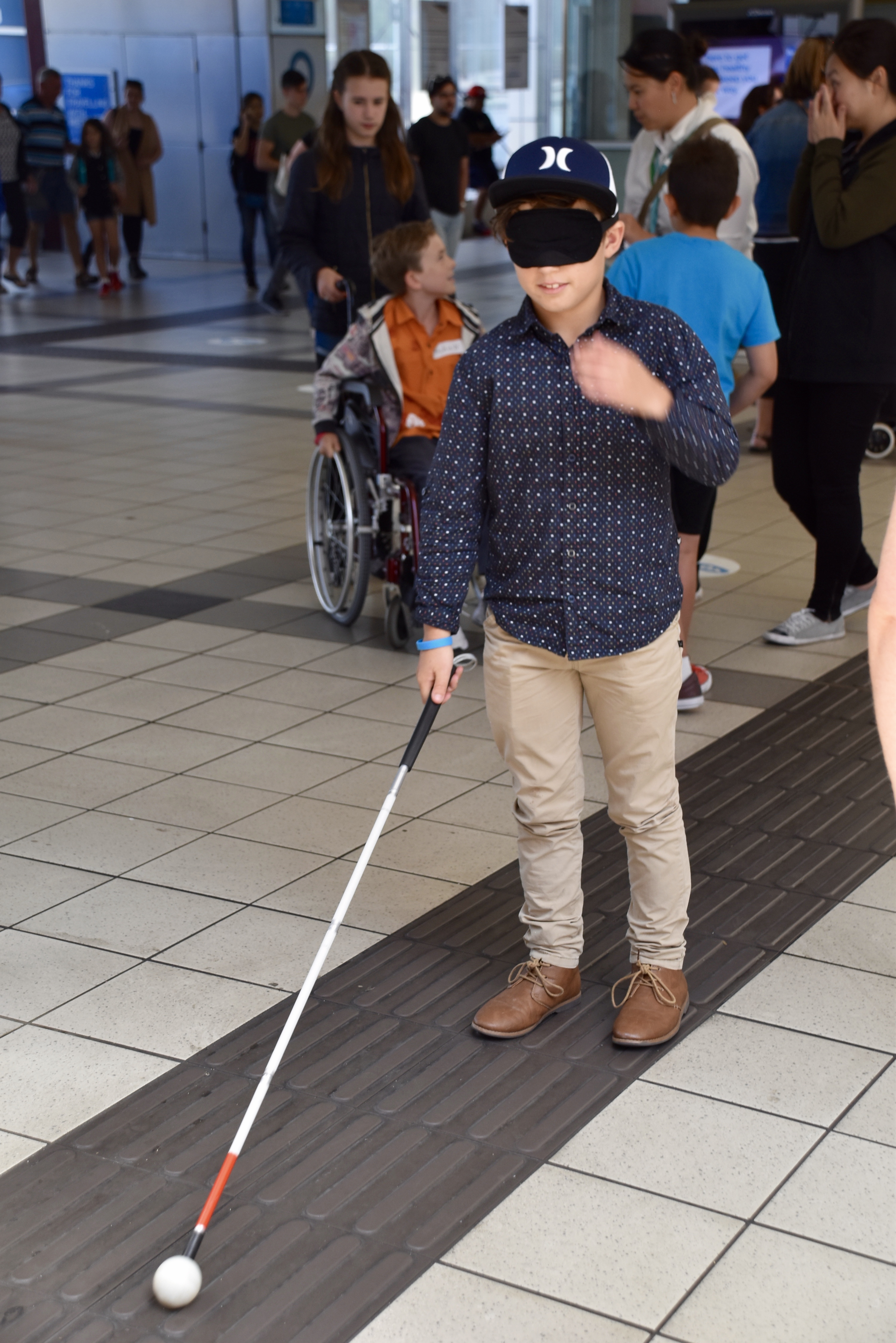 A child of a Public Transport Victoria employee is bl;indfolded and is using a long white cane to navigate through a railway station.