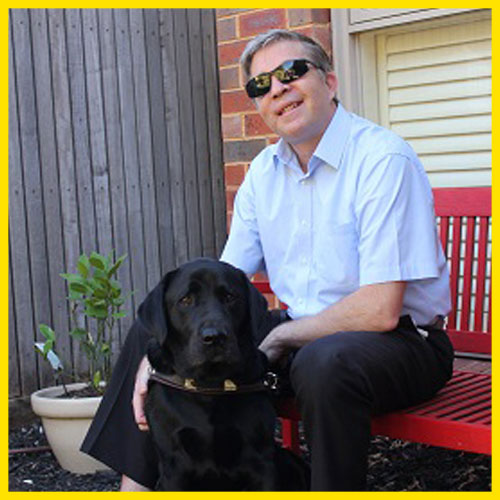 Hamish is sitting on a chair alongside his Seeing Eye Dog