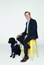 VA_Chris+Edwards_0066 (1) Chris Edwards sitting on a yellow chair next to Odie his Seeing Eye Dog