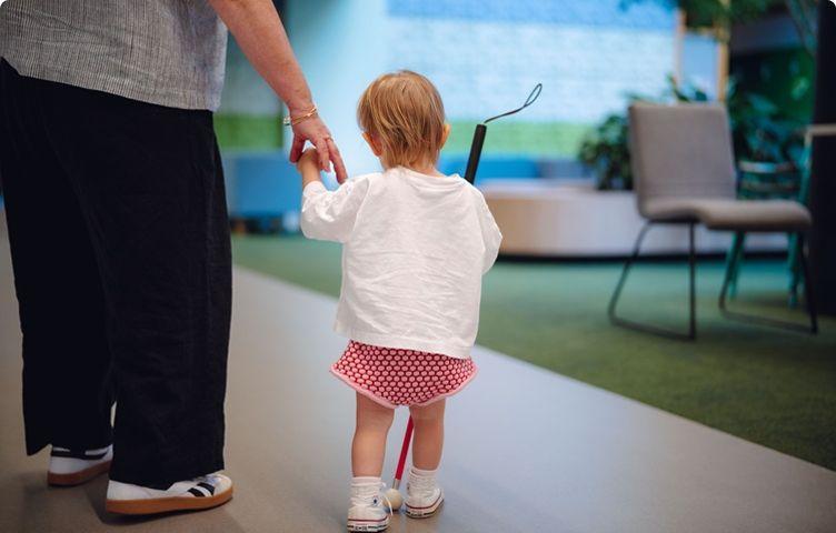 A young child is walking, holding a white cane and a Vision Australia team member's hand.