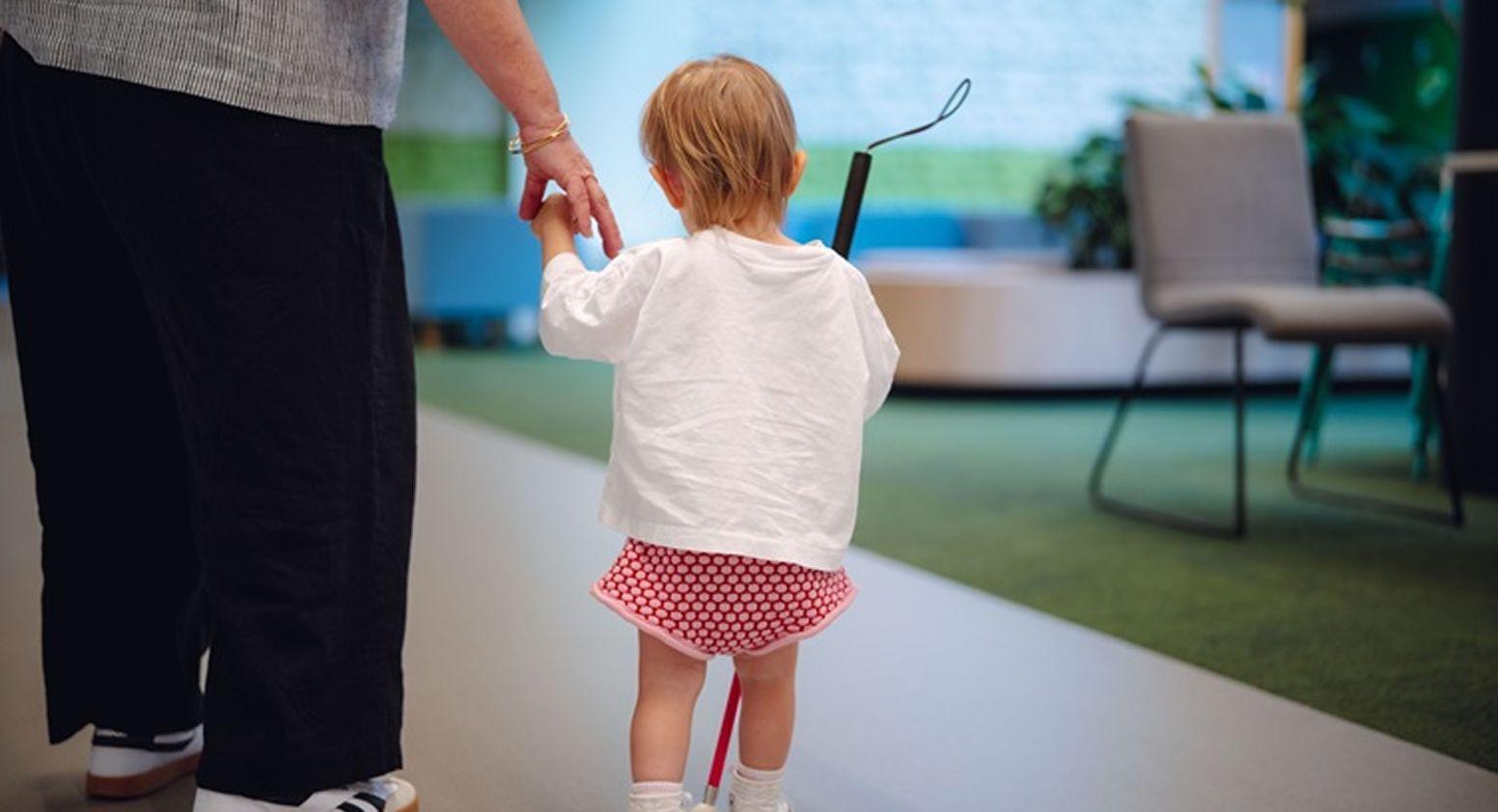 A young child is walking, holding a white cane and a Vision Australia team member's hand.