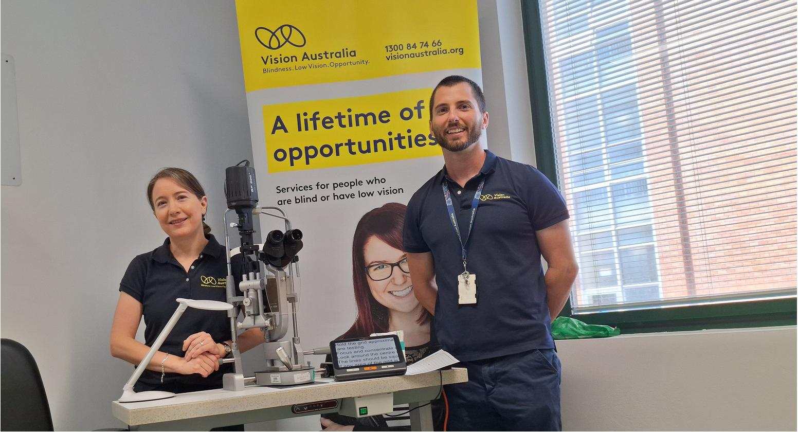 A Vision Australia optometrist and occupational therapist are smiling and standing behind a desk with assistive technology at the Know Your Vision clinic at the Sydney Eye Hospital.