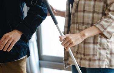 A close up image of two people walking together. One of the people has a white cane and is holding the elbow of the other person.