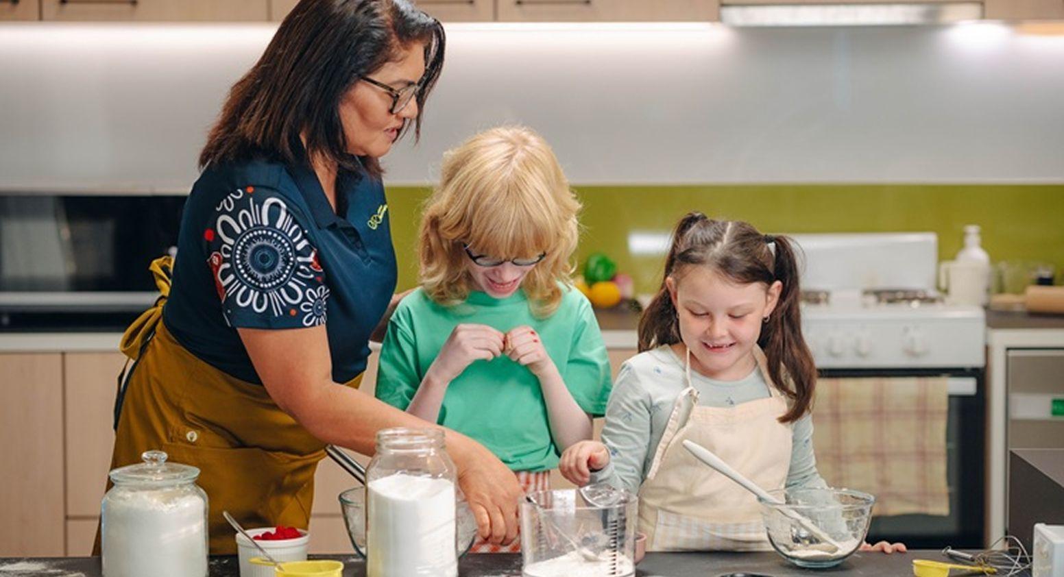 A Vision Australia team member is helping two children cooking in a kitchen.