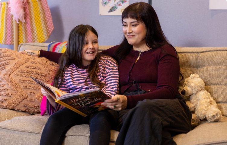 A mum and her daughter with low vision sit on the couch reading a book together.