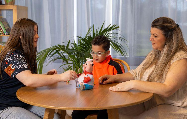 A mum, her son who has low vision and their service provider play an accessible game at a dining table.