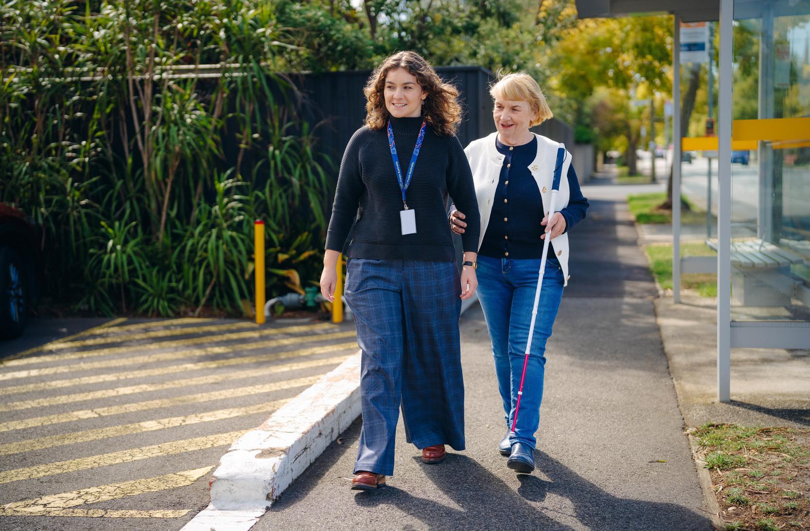 Two women are walking together down the street, one of the women has a white cane.