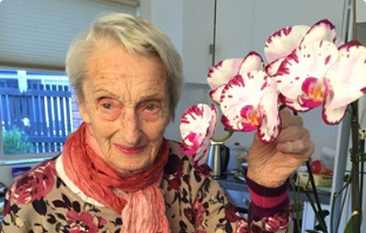 A woman is smiling, she is wearing a scarf, a floral top, she has grey hair and is holding a stem of white and pink orchid flowers.